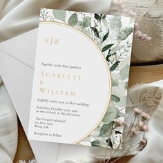 a white and gold wedding card with greenery on the front is surrounded by flowers