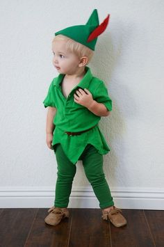 a little boy dressed up in a green outfit and red hat, standing against a white wall