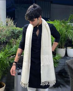 a man in black shirt and white scarf standing next to potted plants with one hand on his hip