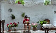 a woman sitting in a chair reading a book next to potted plants and flowers