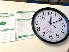 a black and white clock sitting on top of a table next to a spiral notebook