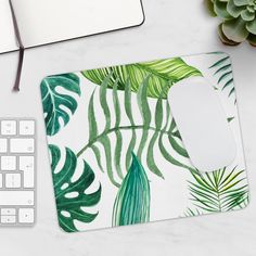 a computer mouse pad with green tropical leaves on it next to a keyboard and plant