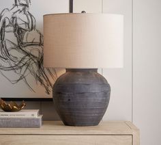 a table lamp sitting on top of a wooden dresser next to a painting and a book