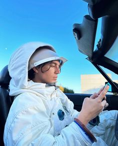 a person sitting in a car using a cell phone while wearing a white jacket and hood