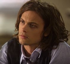 a man with long hair wearing a vest and looking off to the side while sitting down