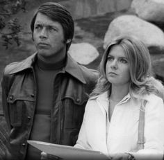 a man and woman standing next to each other in front of a rock wall holding papers