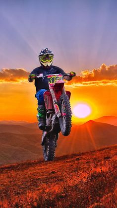 a man riding a dirt bike on top of a grass covered hillside at sunset with the sun in the background