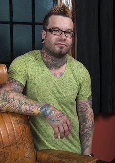 a tattooed man sitting in a chair with his arms crossed and looking at the camera