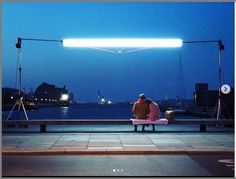 two people sitting on a bench in front of a body of water with lights above them