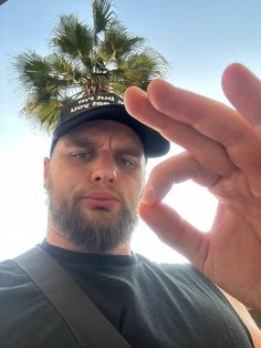 a man wearing a hat and holding his hand up to the camera with palm trees in the background