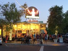 an ice cream shop is lit up at night with people standing outside and on the sidewalk