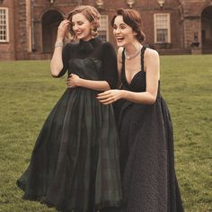 two women in black dresses standing next to each other on a grass covered field with an old brick building behind them