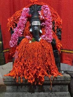 the statue is decorated with flowers and garlands on it's head, sitting in front of a red curtain