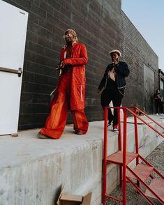 two people are walking down the sidewalk in front of a building, one is wearing an orange outfit