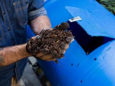 a man holding soil in his hand near a blue barrel filled with composting material