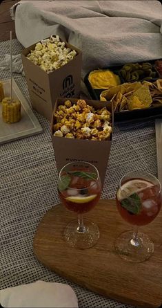 two wine glasses filled with drinks sitting on top of a wooden table next to snacks