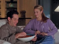 a man and woman sitting on a couch eating food from a white bowl in front of them