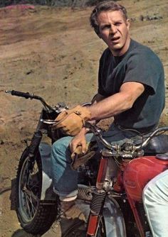 a man sitting on top of a red motorcycle in the middle of dirt road next to trees