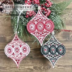 three ornaments hanging from a christmas tree on a wooden table with pine cones and berries in the background