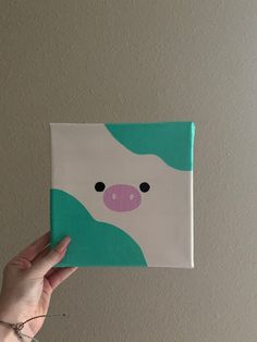 a hand holding up a small square painting with a pig on it's face