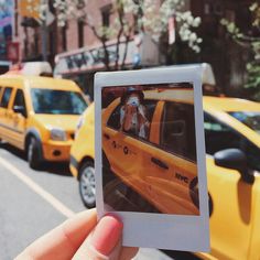 a person holding up a polaroid photo in front of taxi cabs on a city street