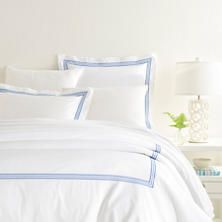 a bed with white sheets and blue trimmings in a bright room, next to a lamp