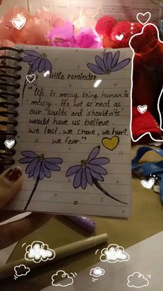 a notepad with writing on it in front of flowers and hearts, next to a hand holding a pen