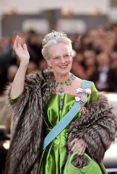 an older woman in a green dress and fur stoler waves to the crowd as she walks