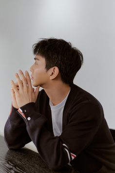 a young man sitting at a table with his hands clasped up to his chest and looking off into the distance