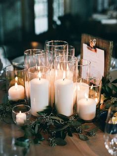 a table topped with lots of white candles next to glasses filled with water and greenery