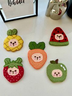 four small knitted fruits and vegetables on a white table with a sign in the background