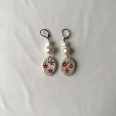 two white and red earrings with pearls hanging from hooks on a white surface, one is decorated with flowers