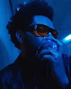 a man wearing sunglasses drinking from a glass in the dark with blue light behind him