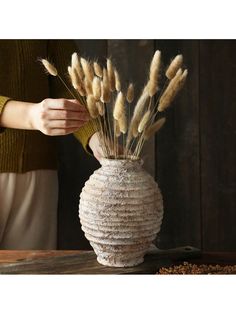 a woman is arranging dried flowers in a white vase on a wooden table next to a wall