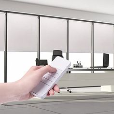 a hand is holding a remote control in an office setting with windows and desks