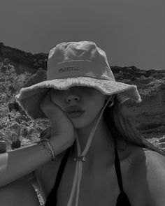 a woman wearing a hat on top of a rocky hill next to the ocean with her hand under her face