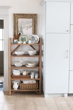 a wooden shelf filled with plates and bowls on top of a hard wood floor next to a white cabinet