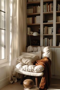 a white chair sitting in front of a window next to a book shelf filled with books