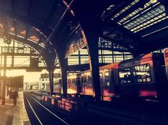 a train station with two trains parked on the tracks and people walking near by at sunset