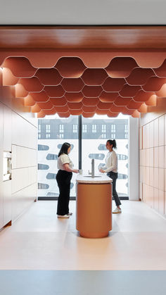 two women are standing in an office with orange and white accents on the ceiling, while one woman stands at a counter