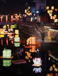 lanterns floating on the water at night with people standing near by and looking at them