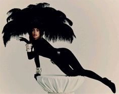 a woman in a black bodysuit and ostrich feathers on top of a vase