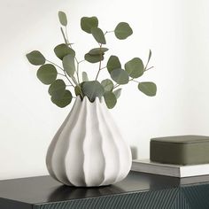 a white vase filled with green leaves sitting on top of a black table next to a book