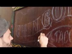 a man writing on a blackboard with white chalk in front of him and wearing a beanie