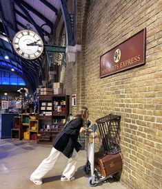 a woman leaning against a brick wall next to a luggage rack with a clock in the background