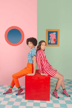 two young women sitting on top of a red box in front of a pink wall