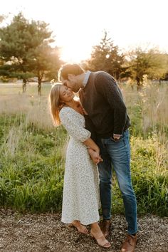 an engaged couple standing in the grass at sunset with their arms around each other and kissing