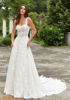 a woman in a white wedding dress standing on a stone walkway with trees and bushes behind her