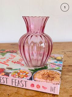 a pink glass vase sitting on top of a wooden table next to a cookbook