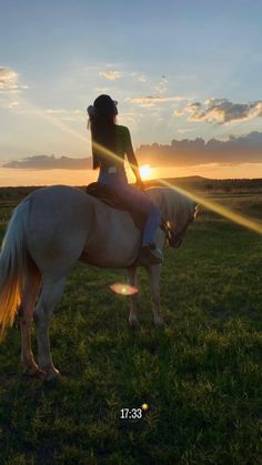 a woman riding on the back of a white horse in a grassy field at sunset